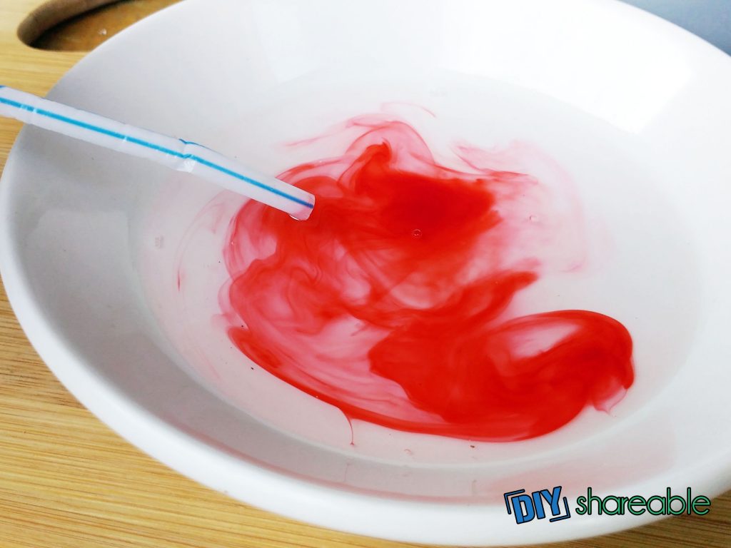 White bowl with a swirl of red food coloring and clear liquid with a blue and white striped straw