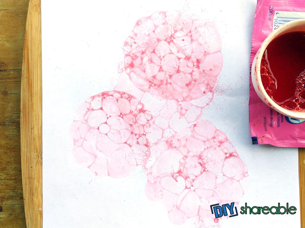 cup of red liquid sitting on top of a white piece of paper with red bubble art print on top of a white table