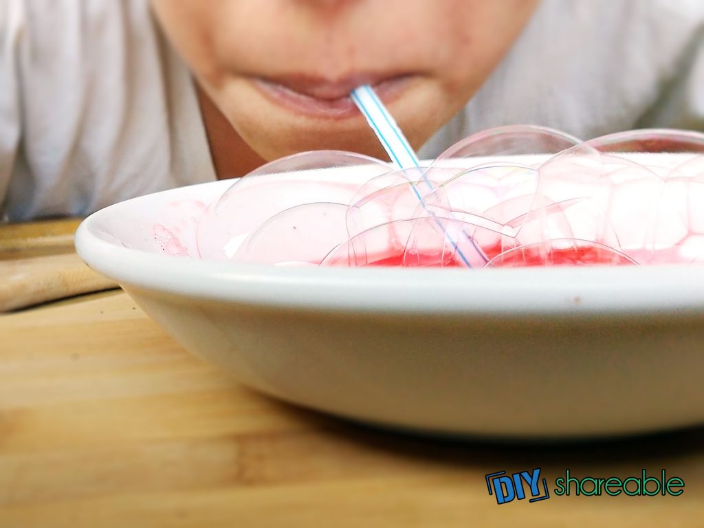 White bowl with red liquid, a person blowing air through a blue and white striped straw with bubbles rising from the dish